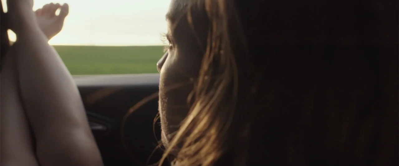 About Us (2017) movie still: closeup — A passenger side interior view of a car with sunlit green fields outside. A woman rests her feet against the…