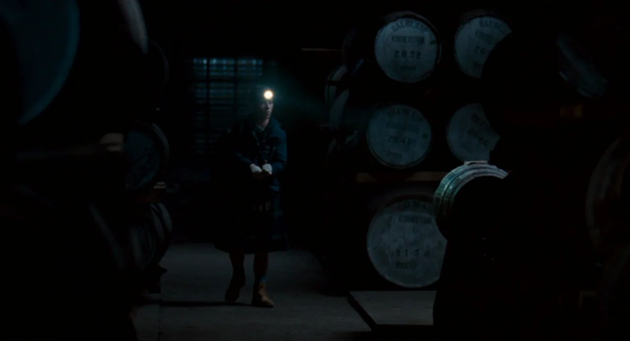 The Angel’s Share (2012) movie still: medium — A dim storage area contains rows of stacked wooden casks marked with white text. A man walks down the…