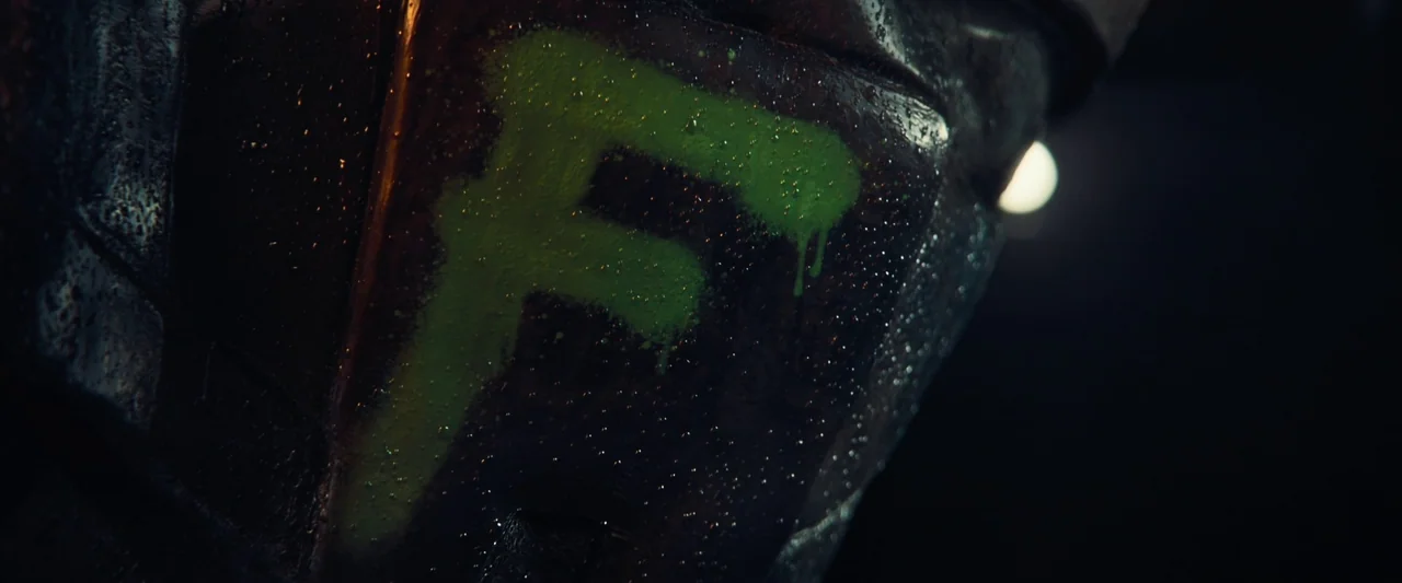 Meanwhile On Earth (2024) movie still: closeup — A dark, wet metallic surface features a large neon green letter F painted in spray. Droplets of…