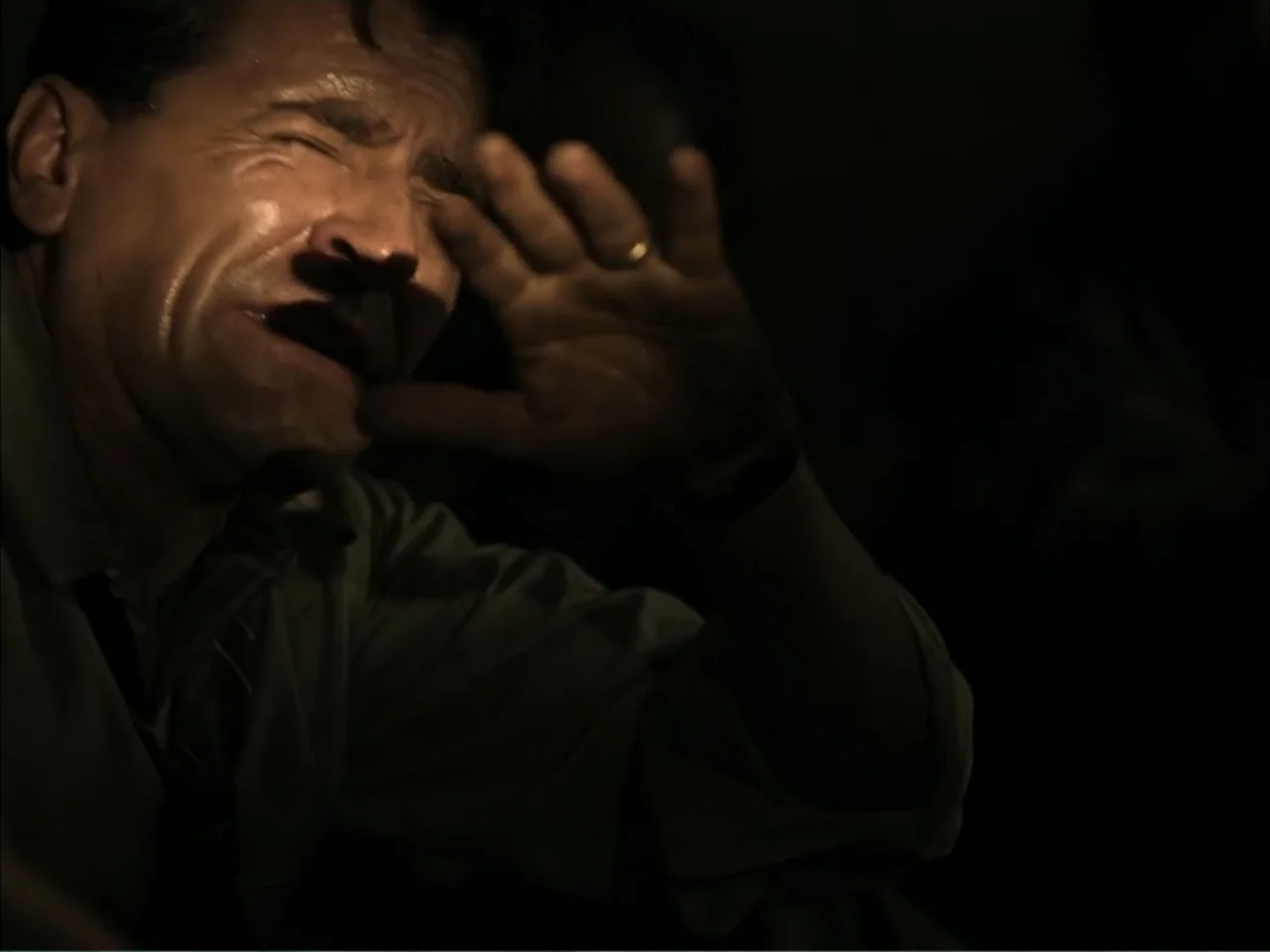 November (2025) movie still: closeup — A dark, dimly lit interior space surrounds a man who appears distressed. The man is holding his hand up in…