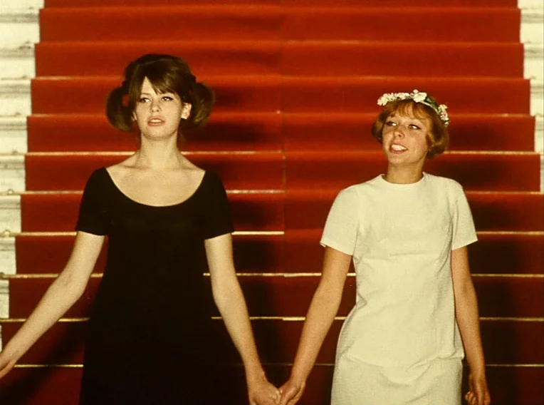 Daisies (1966) movie still: medium — A red-carpeted staircase with white marble side panels serves as the background. Two young women hold hands…