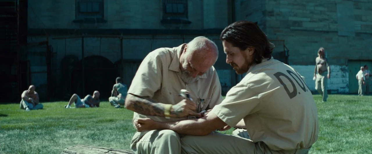 Out of the Furnace (2013) movie still: medium — A prison yard with a large stone wall and a chain-link fence. An elderly man performs a DIY tattoo…