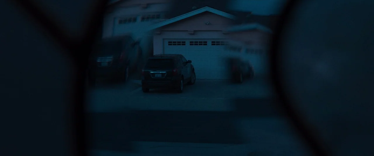 The Perfection (2018) movie still: wide — A suburban house at night seen through a blurred, circular aperture or lens element. A dark SUV is parked…