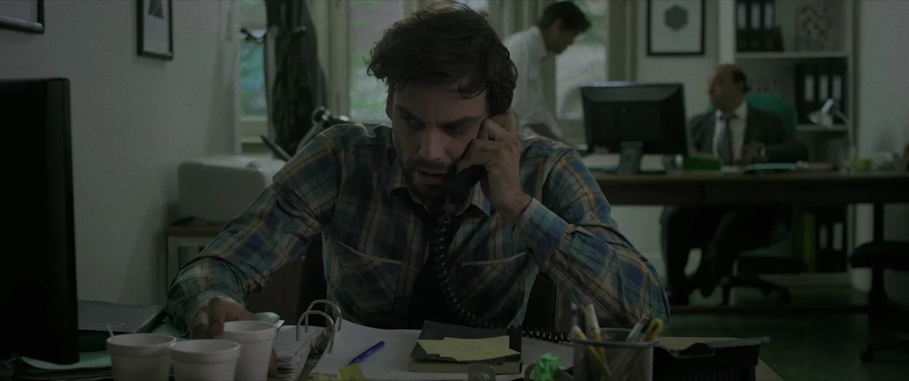 Terrified (2017) movie still: medium — An office environment contains multiple desks separated by partitions and office supplies. A man in a plaid…