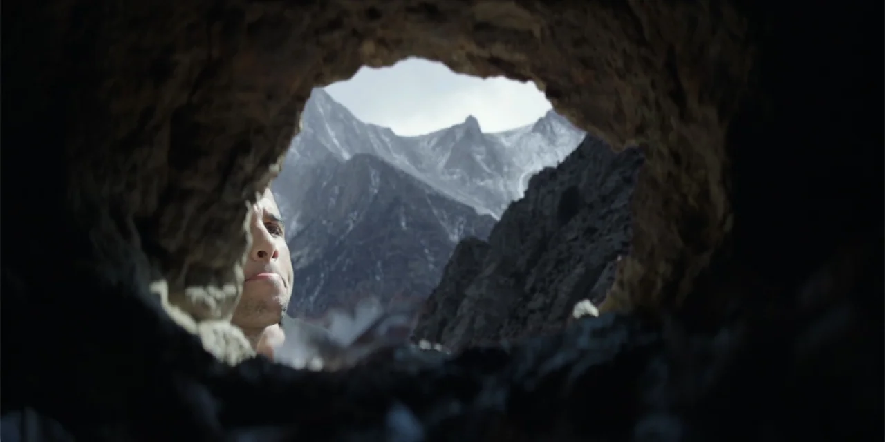 Fall (2022) movie still: medium — A dark, textured cave opening frames a distant view of craggy, snow-dusted mountain peaks. A man stands at the…