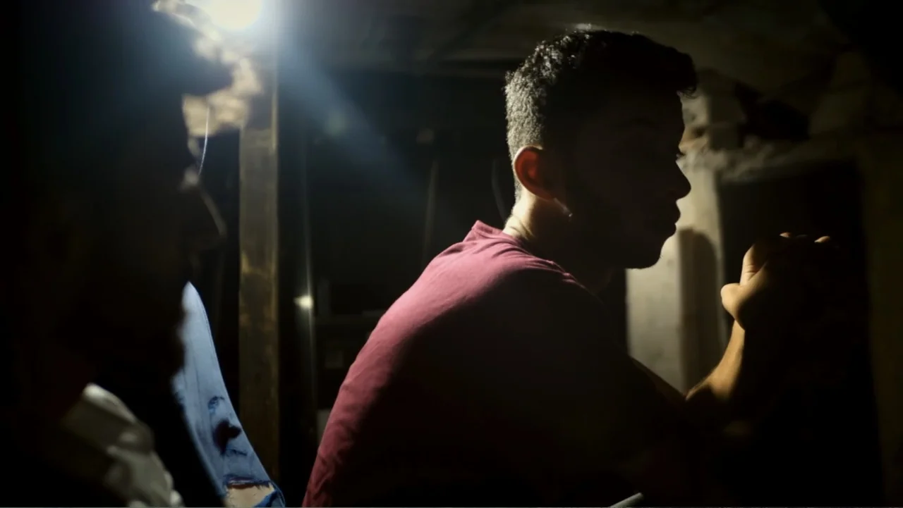 No Other Land (2024) movie still: medium — A dimly lit, unfinished interior space features dark surfaces and low-key directional lighting. Two young…