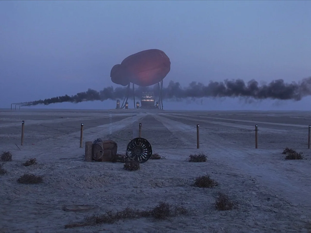 Kin-dza-dza! (1986) movie still: establishing — A flat, dusty desert landscape stretches toward a distant, low-lying industrial facility. A large…