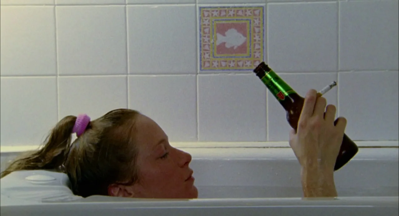 My Summer of Love movie still: medium — A woman rests in a white bathtub tiled with square tiles featuring a fish icon. She holds a green glass beer…