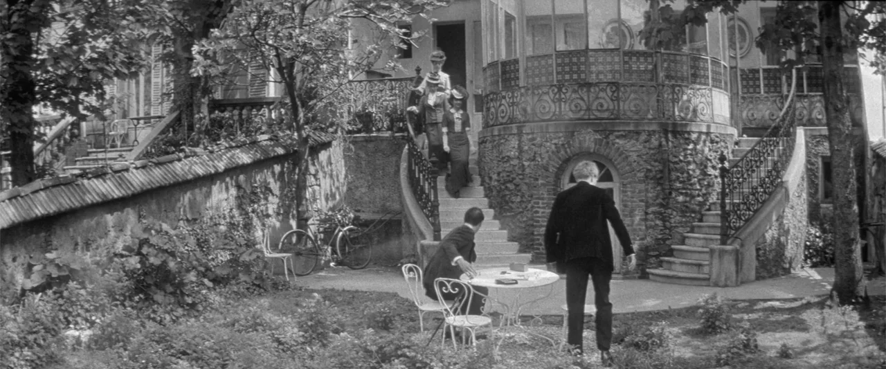 Jules and Jim (1962) movie still: wide — A stone courtyard features an ornate curved staircase and a small outdoor cafe table. Two men stand or sit…