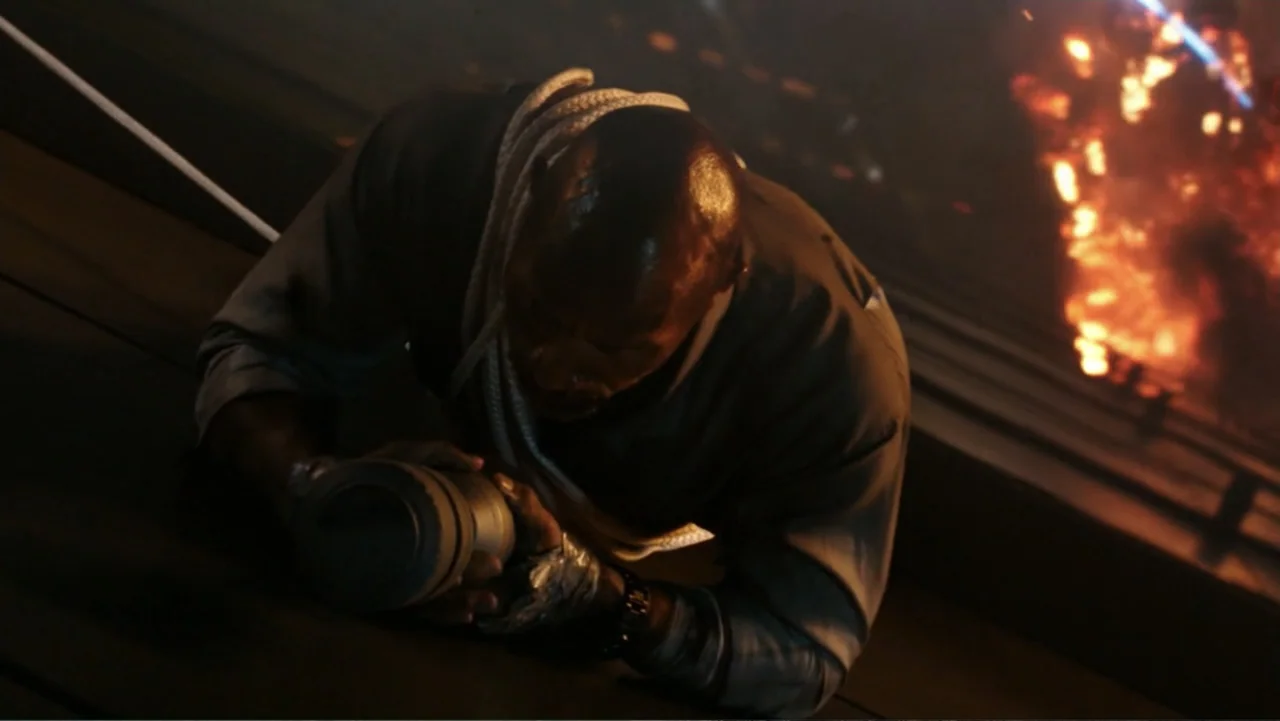Skyscraper (2018) movie still: medium — A dark industrial setting with a bright orange fire burning in the background area. A man crouches in the…