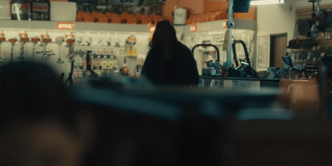 All Roads to Pearla (2019) movie still: medium — A dimly lit retail shop features rows of power tools displayed on orange shelving. A silhouette of…