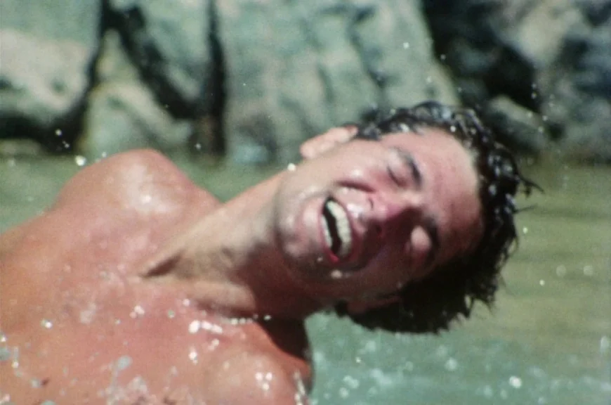 Sebastiane (1976) movie still: closeup — A man stands in shallow water near large wet rocks on a sunny day. He tilts his head back with his mouth…