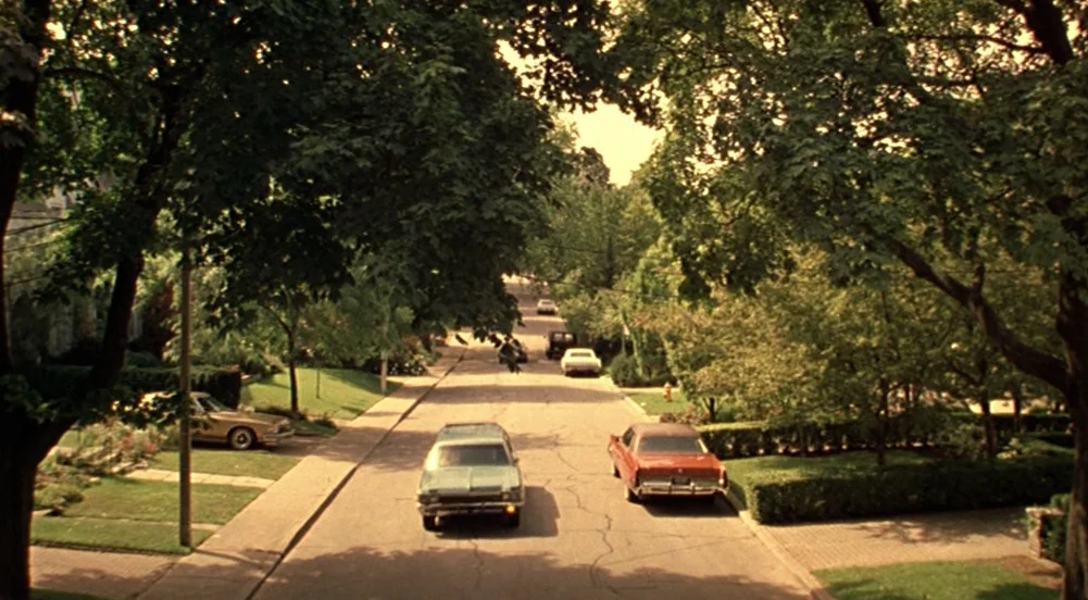 The Virgin Suicides (1999) movie still: wide — A residential street lined with green trees and manicured lawns under a warm sun. Several vintage…