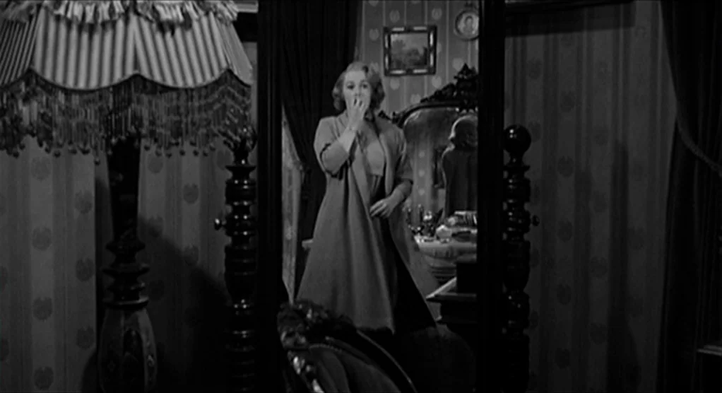 Psycho movie still: medium — A dimly lit room features patterned wallpaper and a large, framed standing vanity mirror. A blonde woman in a coat…