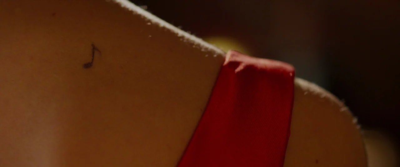 The Perfection (2018) movie still: closeup — A close-up view of a person's shoulder area featuring a small ink tattoo. A red fabric strap rests…