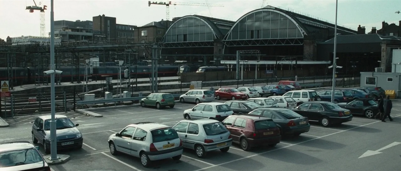 Green Street (2005) movie still: establishing — A paved parking lot sits adjacent to a large railway station with arched roofs. Numerous passenger…