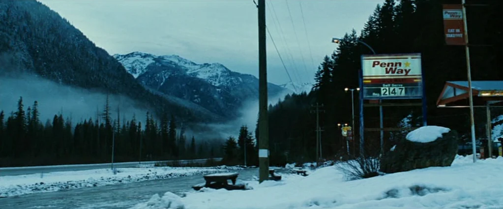 Wind Chill (2007) movie still: wide — A desolate, snow-dusted roadside features a Penn Way gas station sign near a mountain. A river runs along the…