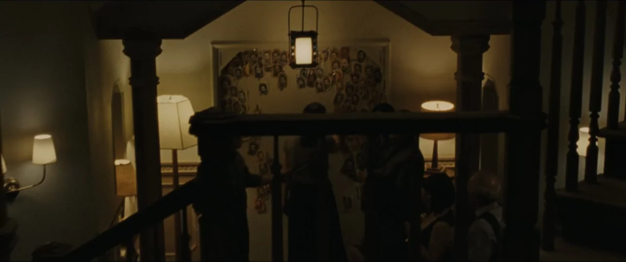mother! (2017) movie still: medium — A dimly lit hallway features a wall covered with numerous taped-up photographs. Several individuals stand in…