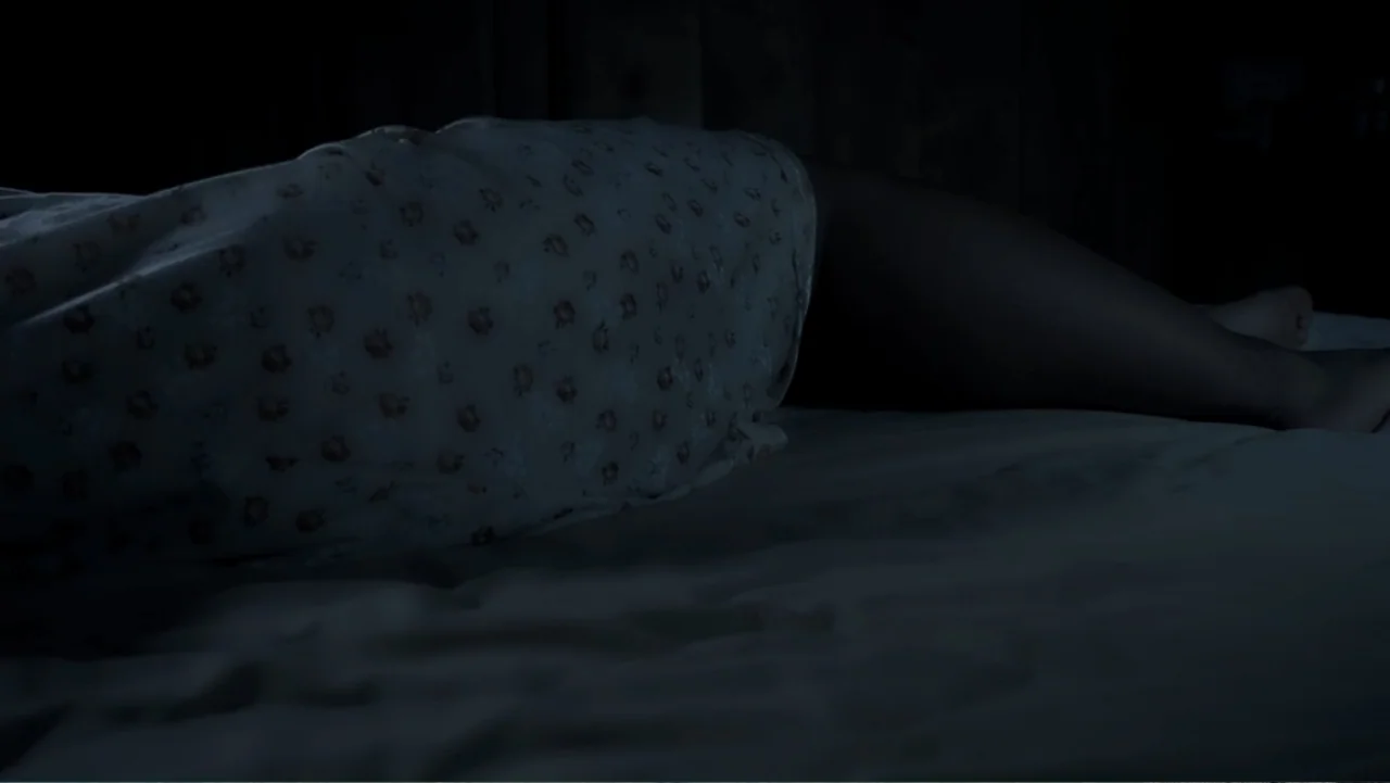 El Duende Sátiro (2016) movie still: medium — A dark bedroom interior shows a person lying under white floral patterned bedding. The subject is…