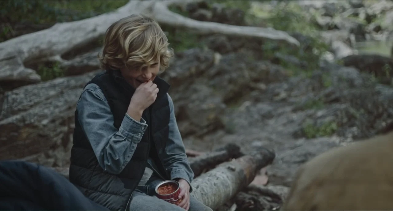 Cowboys (2014) movie still: medium — A muddy riverbank with fallen logs and scattered rocks sits in the outdoors. A young blond boy sits on a log…