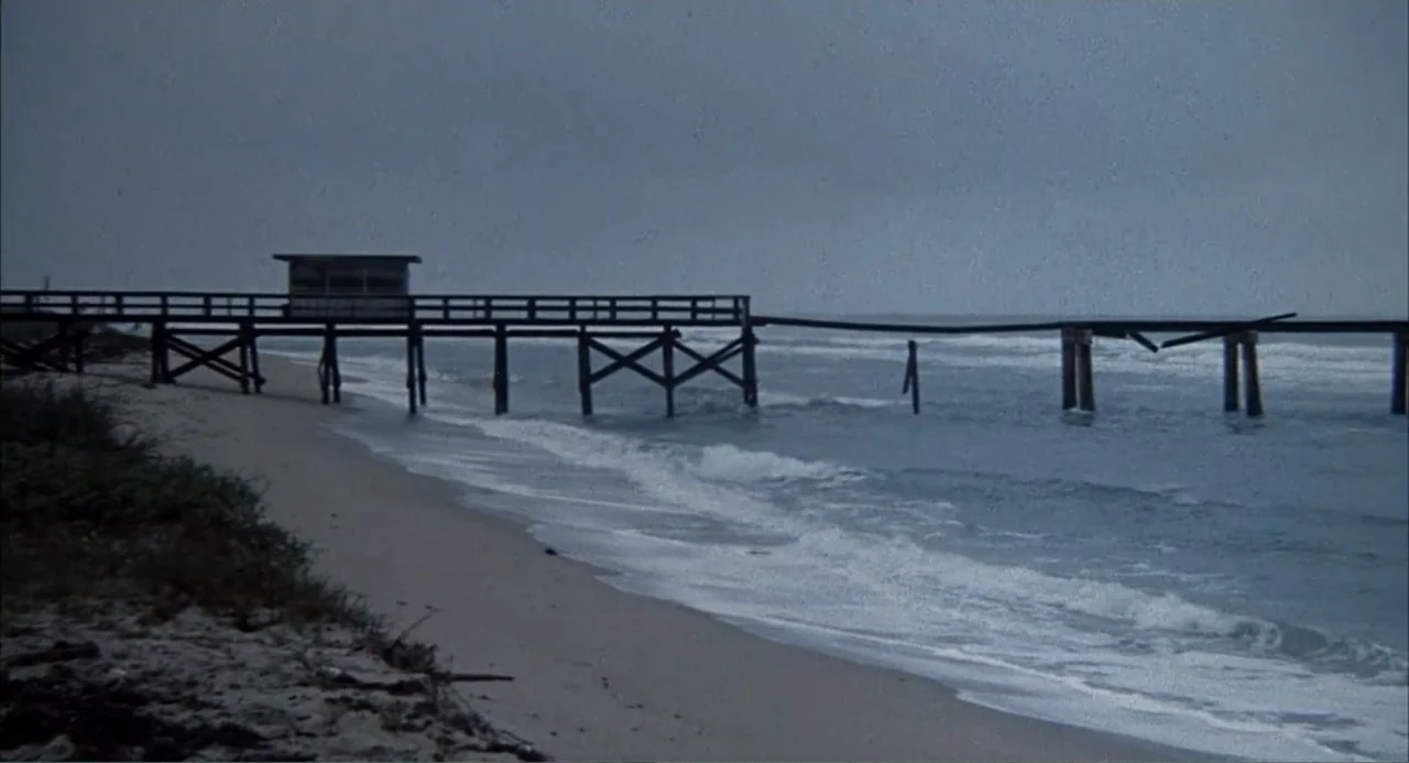 Empire of the Ants (1977) movie still: wide — A long wooden pier stands above the ocean surf on a grey, overcast day. The structure is damaged with…