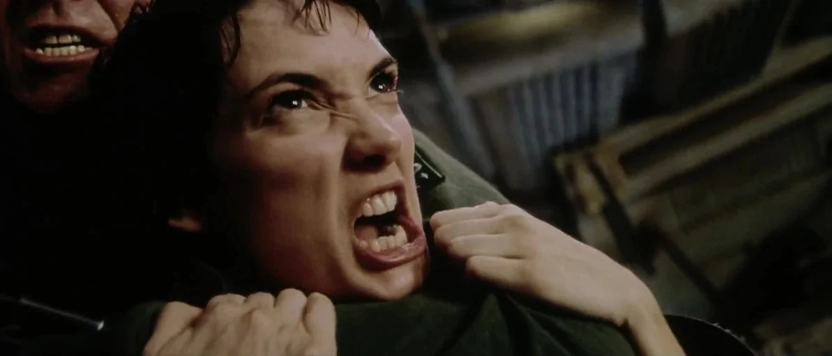 Alien: Resurrection (1997) movie still: closeup — A dark, cramped interior space of a spaceship with metallic structures visible in shadow. A young…