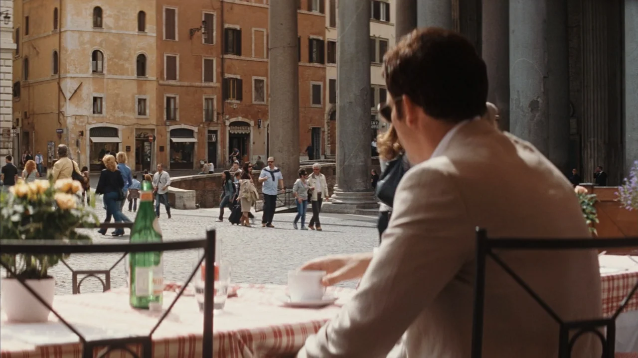 Duplicity (2009) movie still: medium — An outdoor cafe table overlooks a busy cobblestone piazza in front of ancient stone columns. A man in a tan…