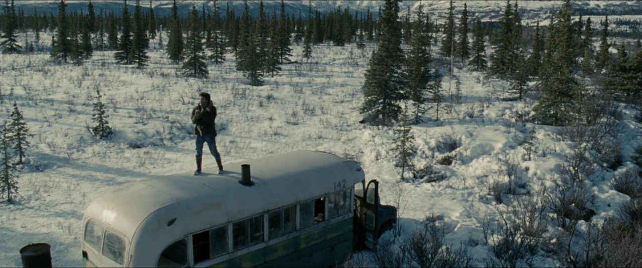 Into the Wild (2007) movie still: wide — A snow-covered clearing with scattered spruce trees surrounds a decaying, abandoned vintage bus. A lone man…
