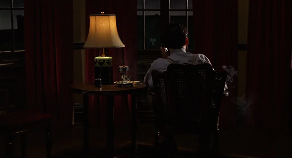 Miller’s Crossing (1990) movie still: medium — A dimly lit room at night features heavy red curtains against dark windows. A man sits in a wooden…
