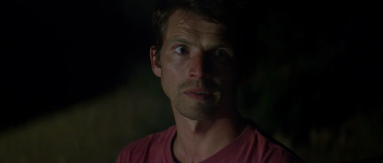 Stranger By The Lake (2013) movie still: closeup — A man stands outdoors at night with a dark, indistinct background behind him. He gazes off-frame…