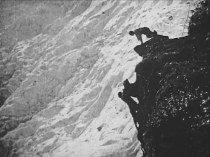 A vertical rock cliff side sits beside a vast, snow-covered glacier slope. Two climbers scale the dark, sheer rock face using traditional mountaineering methods.