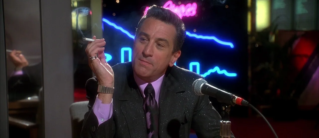 Casino (1995) movie still: medium — A dark broadcast studio features a neon sign glowing against a black backdrop. A man in a suit sits before a…