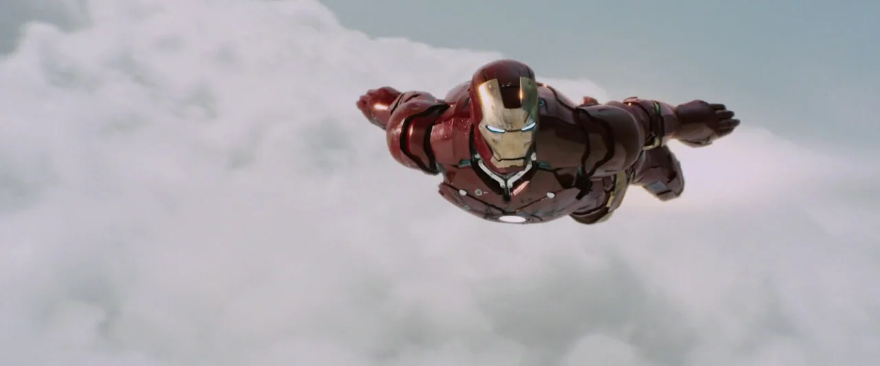 Iron Man (2008) movie still: medium — A high-altitude aerial view features clouds surrounding a figure in armored metallic gear. The figure flies…