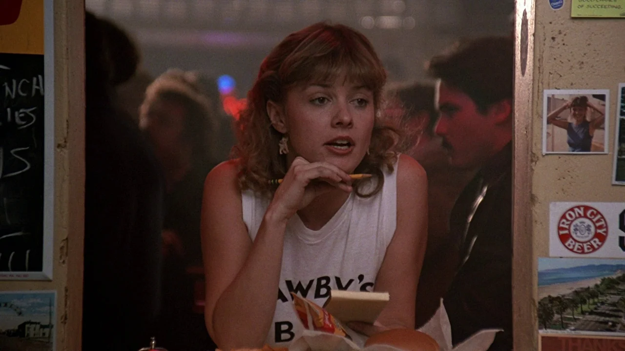 Flashdance (1983) movie still: medium — A cluttered wall featuring photographs and beer stickers frames a woman in a booth. The woman wears a white…