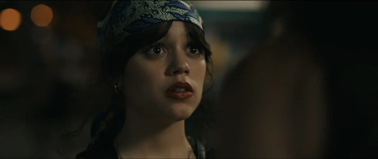 Scream VI (2023) movie still: closeup — A nighttime exterior street scene with dark surroundings and blurred lights. A young woman with a headscarf…