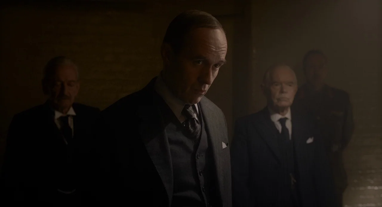 Darkest Hour (2017) movie still: medium — A dimly lit basement features several men in formal suits standing in the shadows. The central man in a…