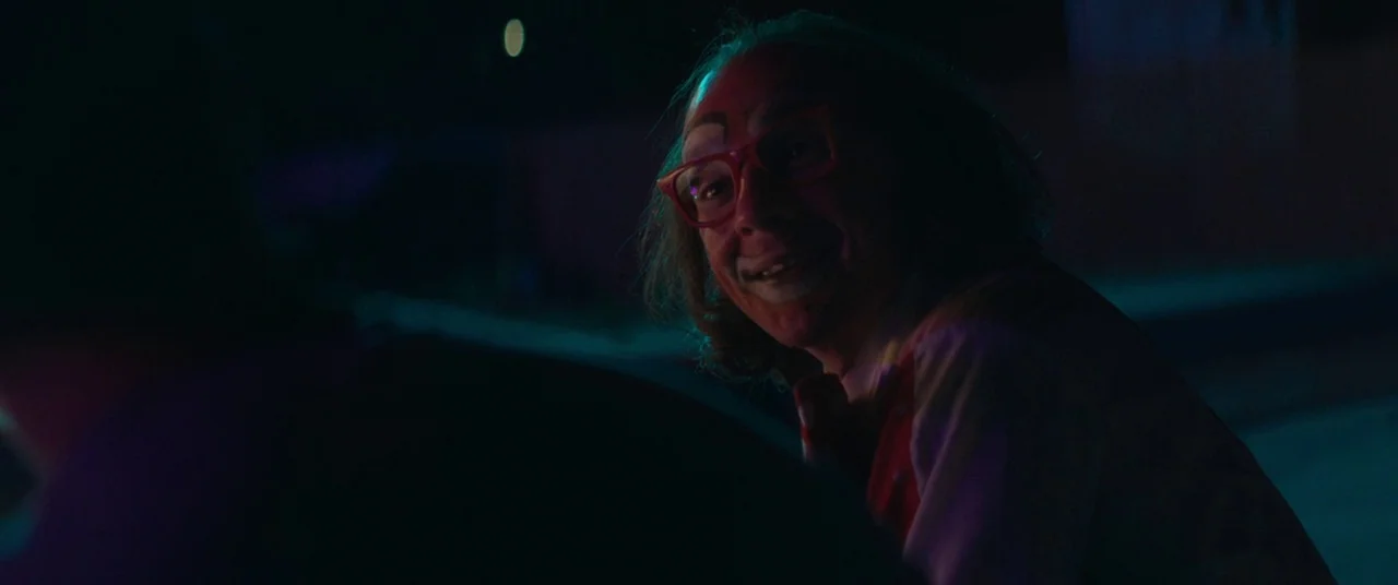 Honey Boy (2019) movie still: closeup — A night exterior setting lit by intense neon lights with a dark foreground silhouette. A man with clown…