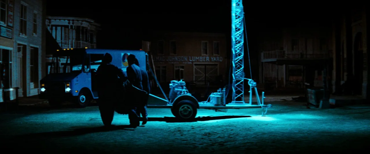 Westworld (1973) movie still: wide — An empty nighttime street features a wooden lumber yard building and a blue van. Two people approach a portable…