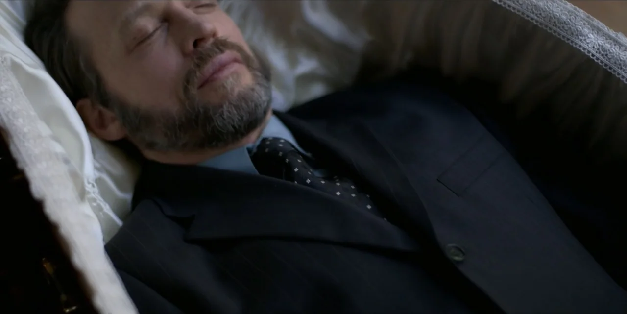 Bring Her Back (2025) movie still: closeup — A man in a formal suit lies inside a satin-lined casket. He is dressed in a dark pinstripe jacket and…