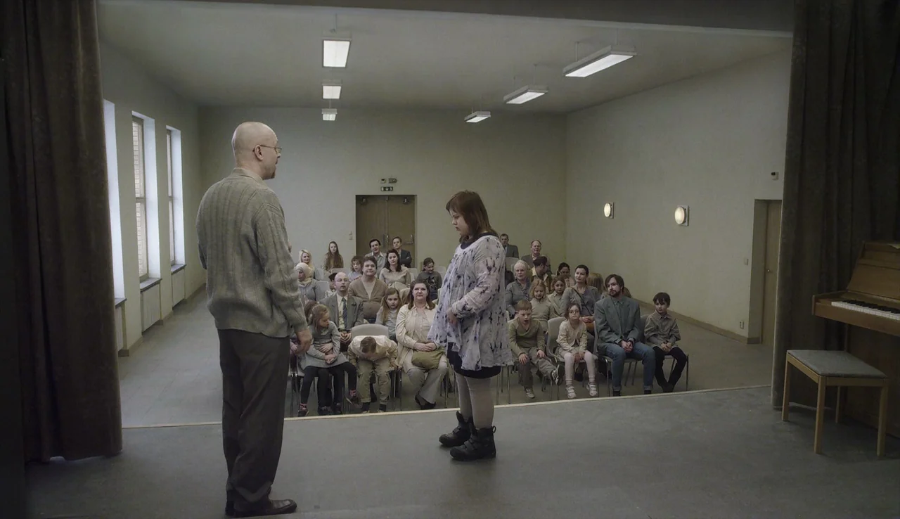 A Pigeon Sat on a Branch Reflecting on Existence (2014) movie still: wide — A sparse community hall with beige walls, rows of chairs, and overhead…