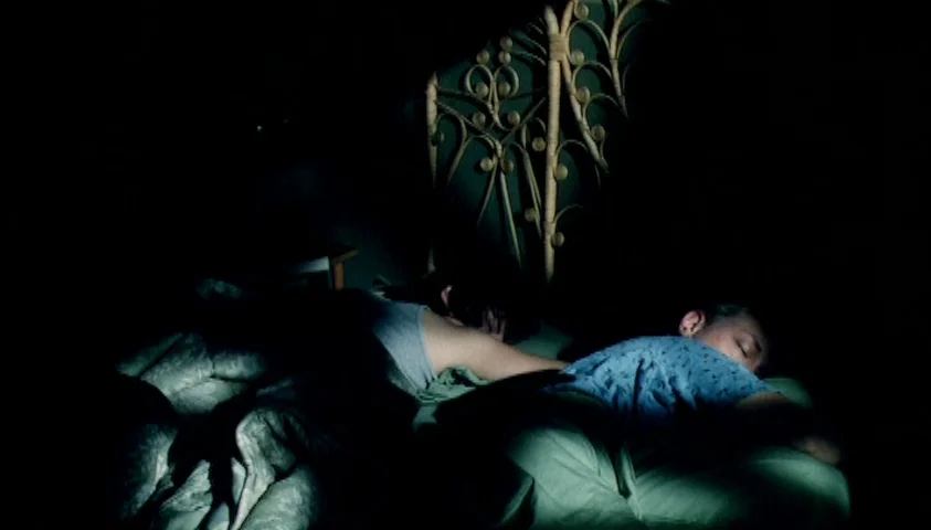 Maelström (2000) movie still: medium — A dark room contains a bed with green sheets and a decorative headboard. Two individuals lie in the bed under…