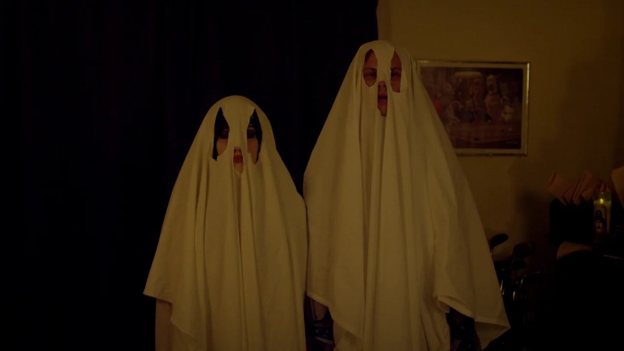Suburban Gothic (2014) movie still: medium — Two figures in a dimly lit room wear white bedsheets as makeshift ghost costumes. They stand side by…