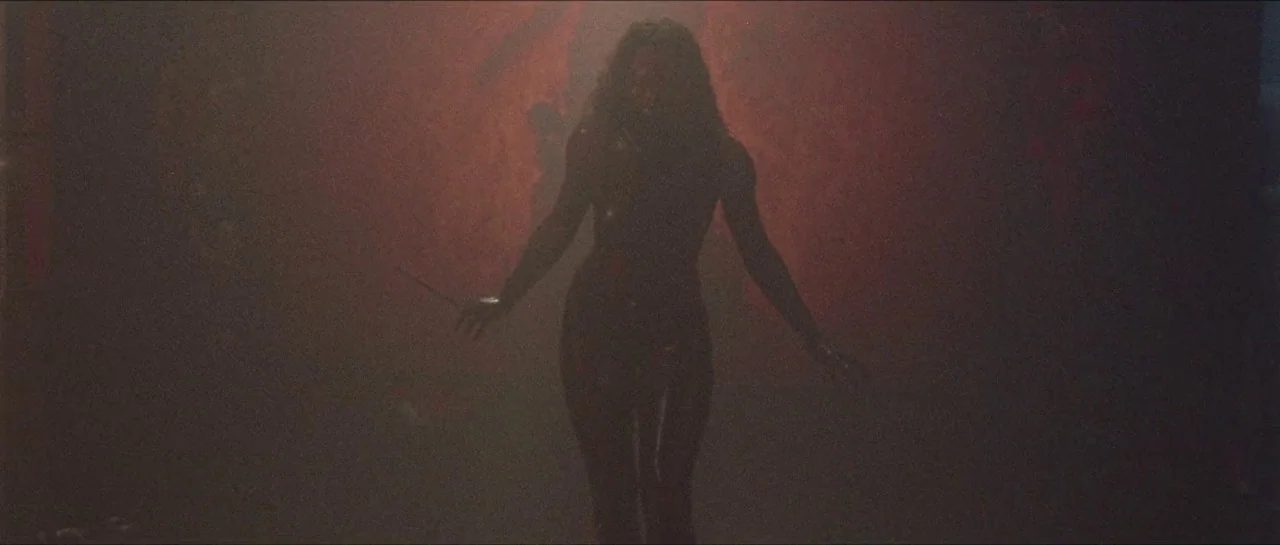 Bliss movie still: fullbody — A dark space lit by a vibrant red glow against a textured backdrop. A woman in dark clothing stands silhouetted…