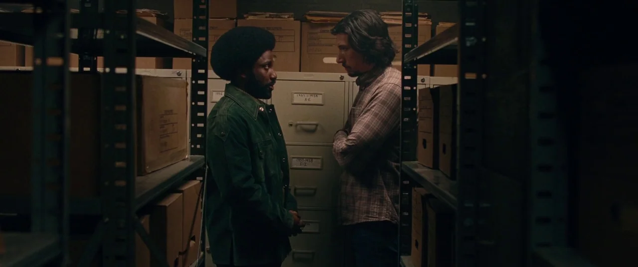 BlacKkKlansman (2018) movie still: medium — A dimly lit archives room contains floor-to-ceiling metal shelving units packed with cardboard boxes.…
