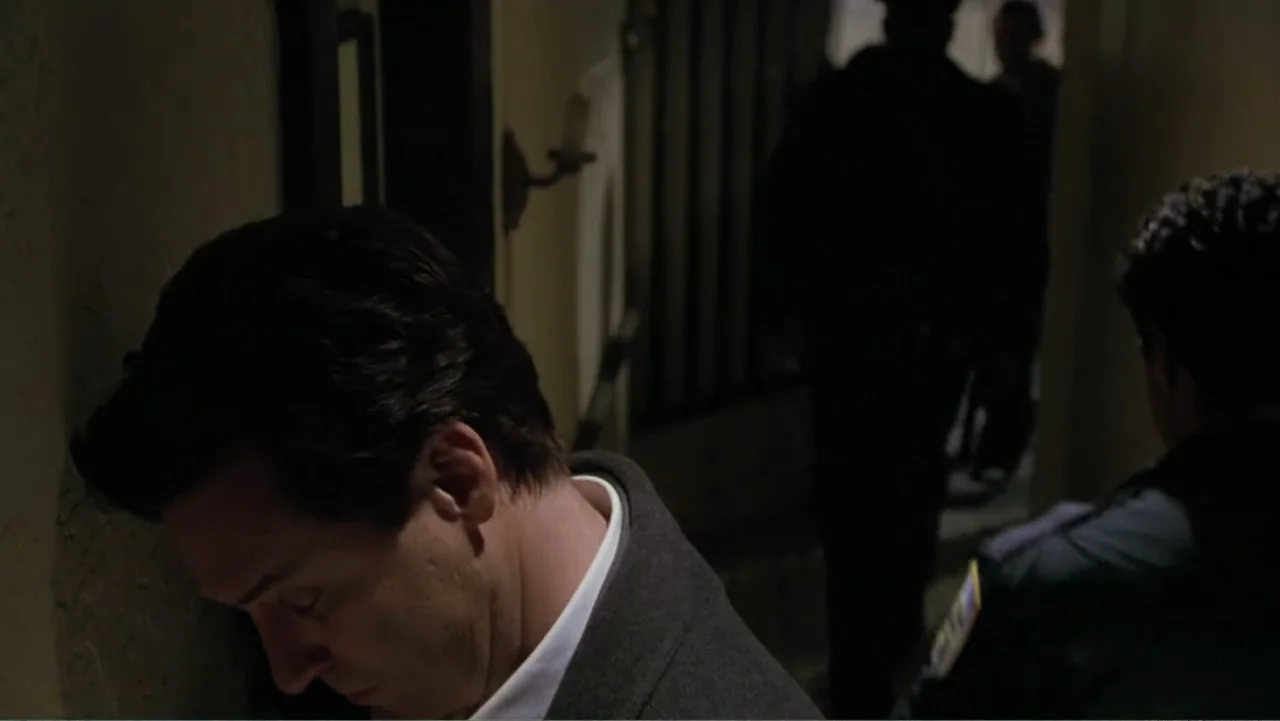 Mistrial (1996) movie still: medium — A dim, narrow indoor staircase features beige plaster walls and dark shadowy recesses. One man rests his head…