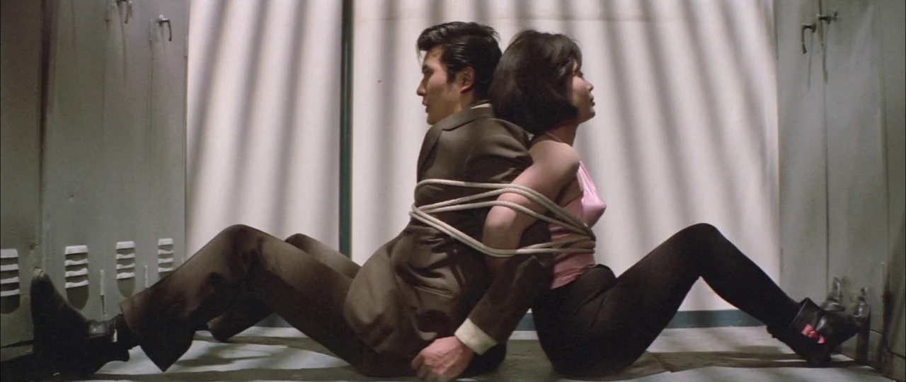 Black Tight Killers (1966) movie still: fullbody — A locker room with beige metal lockers features striped shadows cast across the back wall. A man…