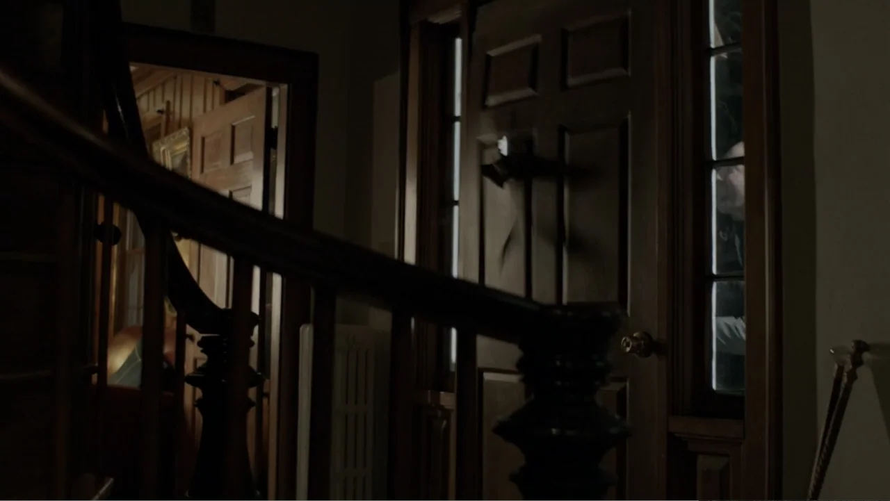 The Collective (2023) movie still: medium — A dark wooden staircase landing features a heavy door with rectangular glass panes. An older man stands…