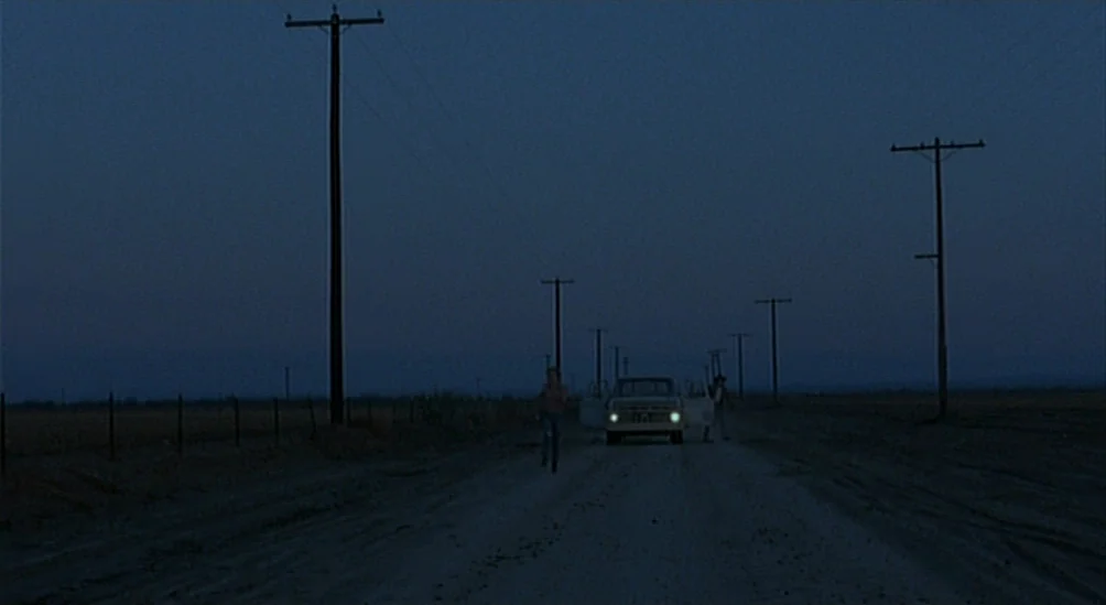 Near Dark (1987) movie still: wide — A dark, unpaved rural road stretches into the distance beneath a vast night sky. A person jogs forward on the…