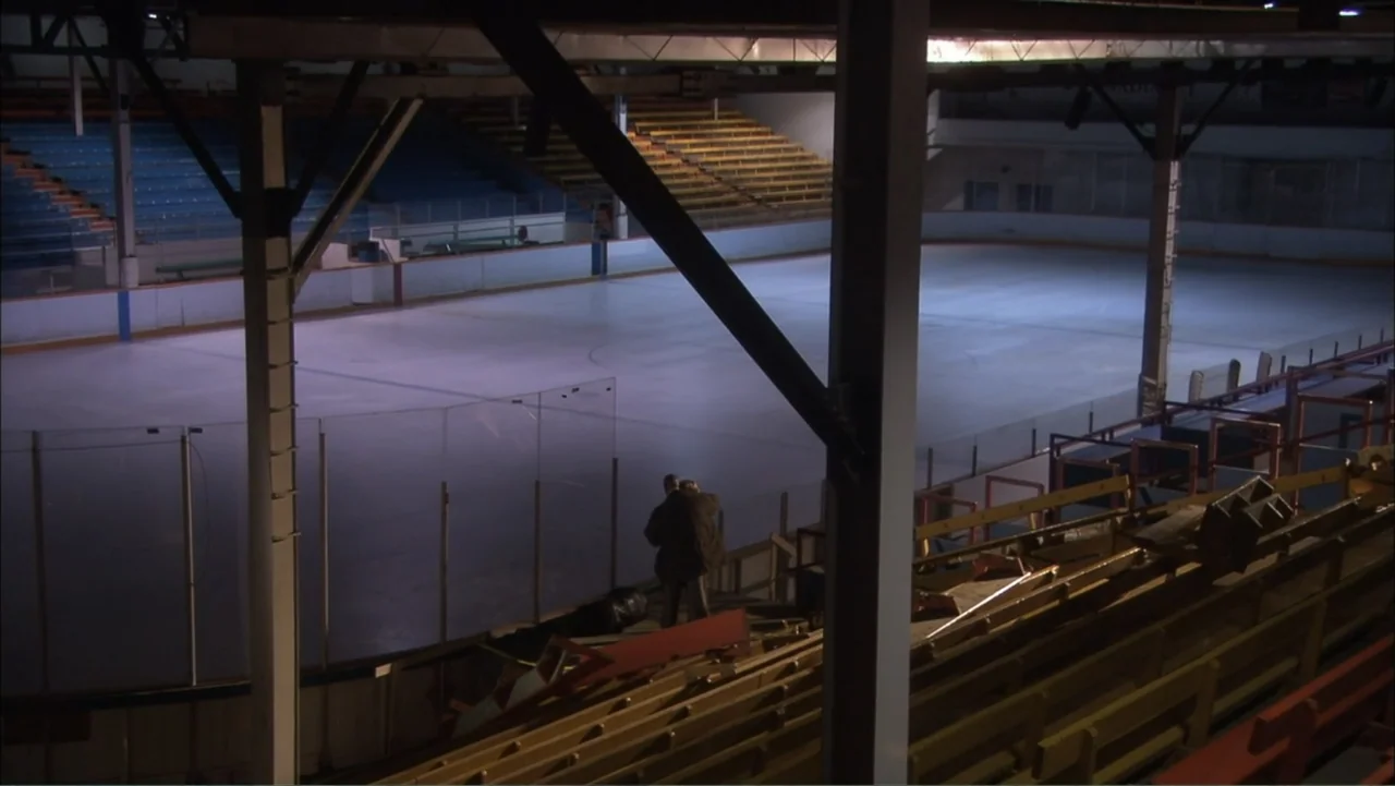 O código oculto (2025) movie still: wide — An indoor ice hockey rink with vacant wooden bleachers and blue plastic seating.