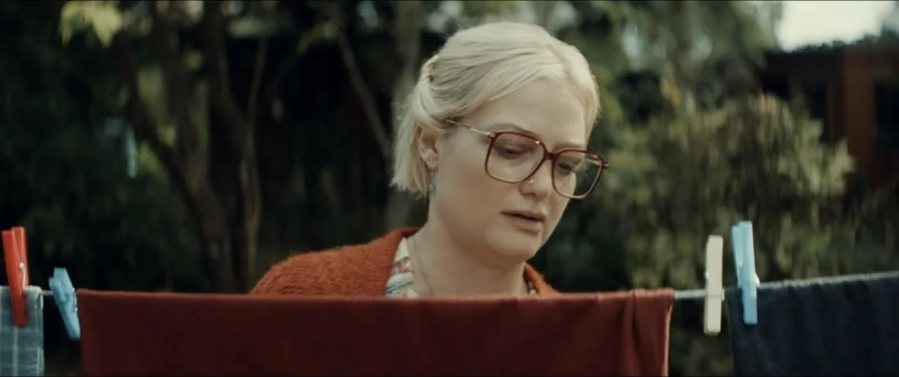 Bonus Track (2024) movie still: medium — A residential backyard with a clothesline running across the foreground. A woman hangs laundry while…